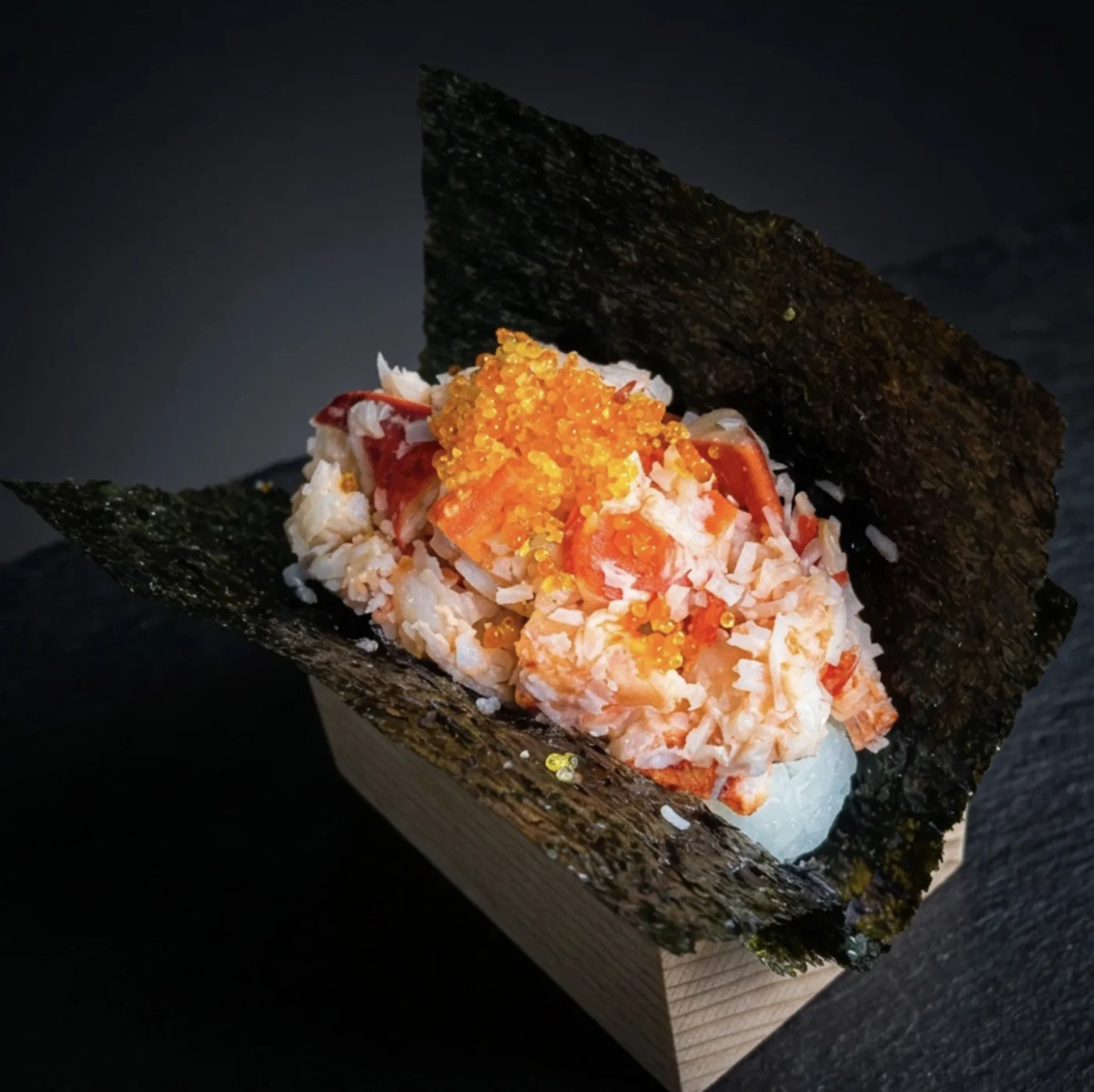 Handroll de lagosta do Kaizen: pedaços de lagosta, arroz e ovas sobre uma folha de nori aberta, servido em pé numa caixa de madeira